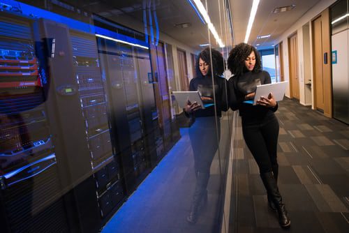 IT woman checking on laptop while leaning on glass room of servers